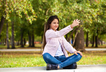 Obraz premium Brunette girl photographs herself on a cell phone while sitting in an autumn park.