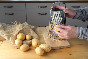 Hands of a woman are grating potatoes with a metal grater on a wooden kitchen table