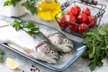 Two raw rainbow trout fish on a plate, greens and fresh vegetables for preparing healthy and tasty food. Healthy diet and delicious culinary concept.
