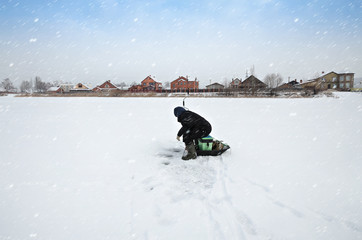 A fisherman sits near an ice hole on a frozen snow-covered river in a snowfall