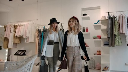 Slow motion, tracking shot of cheerful female friends in fashionable outfit holding shopping bags and going to exit from fashion clothing store satisfied with shopping.