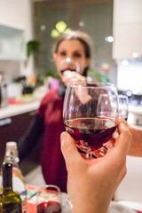 People having fun together toasting drinks in the home kitchen. Happy and smiling woman toasts with glass of red wine. Focus on central glass. Friendship and family concept. Italian wine.
