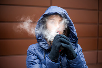 Vape teenager. Young pretty white caucasian girl in a blue jacket and hood smoking an electronic...