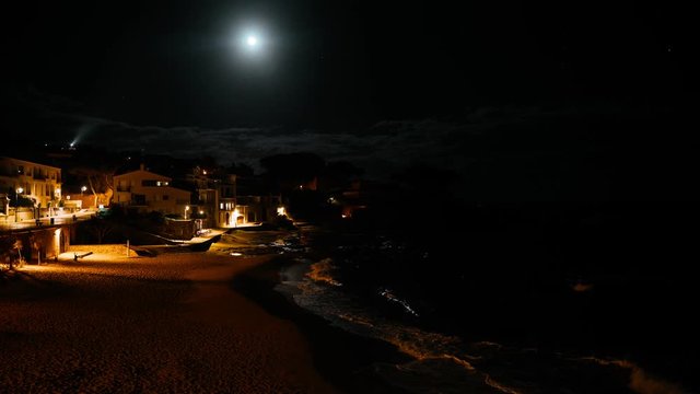 Night view of a small mediterranean fishing village. Fool moon reflection in the water. Night lights on the seashore. Waves splashes in a small bay with a small coastal village. Night sea white houses