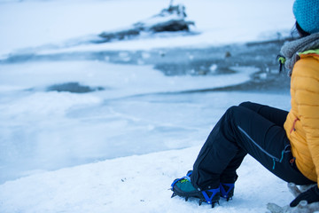 Fototapeta na wymiar Girl sitting by an icy mountain lake with crampons on.yellow jacket, blue hat and black pants.
