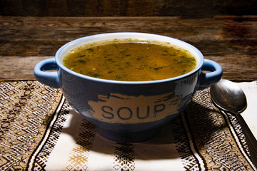Homemade meat soup in a ceramic blue bowl, metal spoon, folklor napkin on a wooden table.