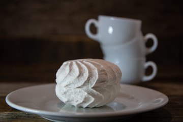 White marshmallows, in a white plate on a wooden table, against the background of white coffee cups.