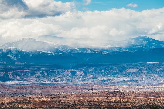 A vast colorful desert landscape under snow-capped mountains and winter storm clouds