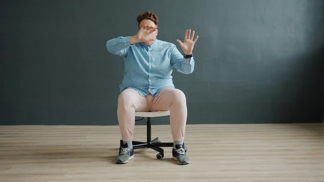 Funny actor is moving arms like touching wall sitting on office chair on dark background having fun making silly faces. Comic gestures and humorous activity concept.