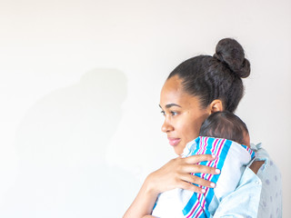 A happy mixed race African American mother wearing a hospital gown holds her new healthy infant baby boy on her shoulder hugging and cradling him as she looks to the side as he sleeps in his swaddle.