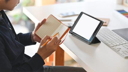 Side shot of the young creative man while writing on the notebook and concentrating to the white blank screen tablet. Keyboard, color guide, notebook and smartphone is on the background.