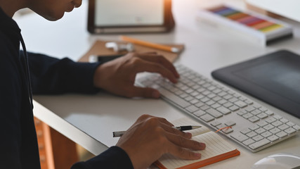 Side shot of young creative man while giving a concentrate on keyboard and notebook at the work desk. Color guide, tablet, earphone, pencil, smartphone on background working desk.