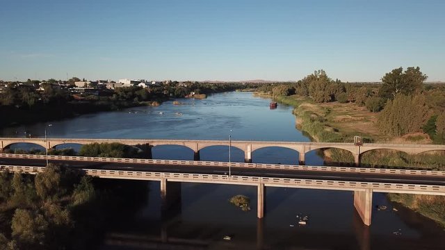 4K summer sunny afternoon aerial drone video: Orange River arched motorway long bridge, grassy islands, boat on the water and busy N10 highway located in town Upington, Northern Cape, South Africa