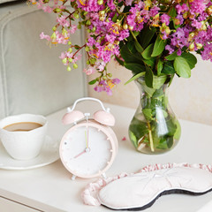 Alarm clock closeup, a cup of coffee and bouquet of pink flowers background in the morning sunlight