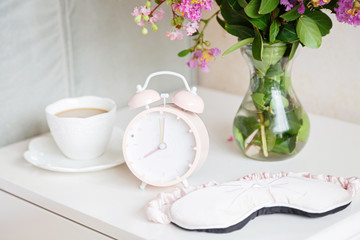 Alarm clock closeup, a cup of coffee and bouquet of pink flowers background in the morning sunlight