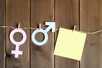 female and male symbols cut out of paper and blank sticky note attached with a clothespin to a rope on a wooden background