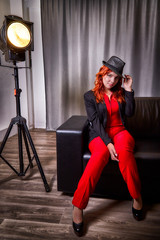 Beautiful young woman with redhair, black hat and red dress on black sofa in the room and nice light behind her