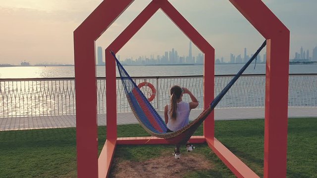 Young woman weared in office sute having coffee in hammock while chilling , freelance life style conceptual, work anywhere. Modern city on the background.