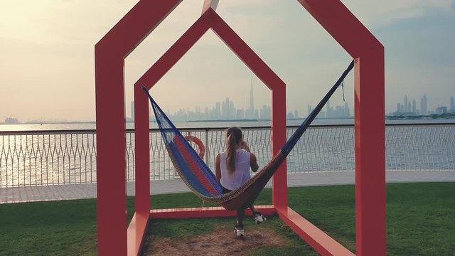 Young woman weared in office sute having coffee in hammock while chilling , freelance life style conceptual, work anywhere. Modern city on the background.