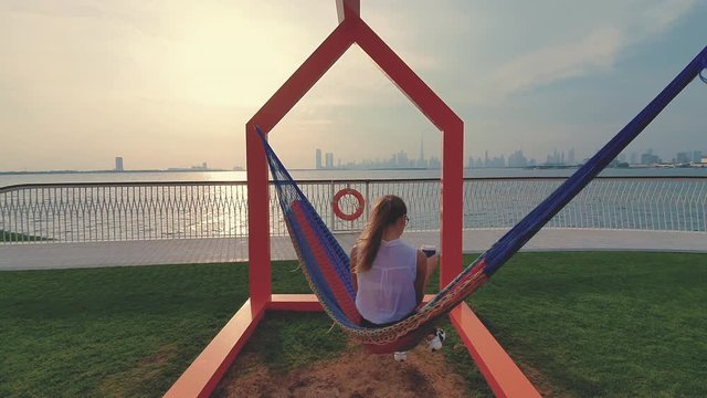 Young woman weared in office sute having coffee in hammock while chilling , freelance life style conceptual, work anywhere. Modern city on the background.