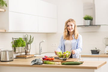 Obraz na płótnie Canvas Young happy blonde girl preparing healthy salad in home kitchen