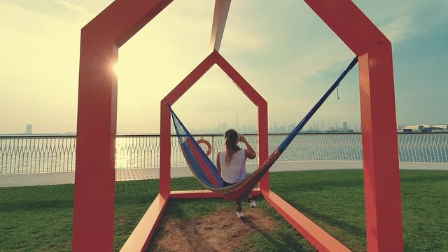 Young woman weared in office sute having coffee in hammock while chilling , freelance life style conceptual, work anywhere. Modern city on the background.