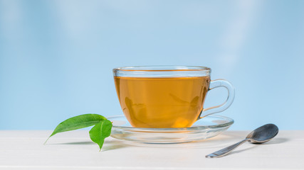 Glass Cup with tea and green leaves on a white table on a blue background.