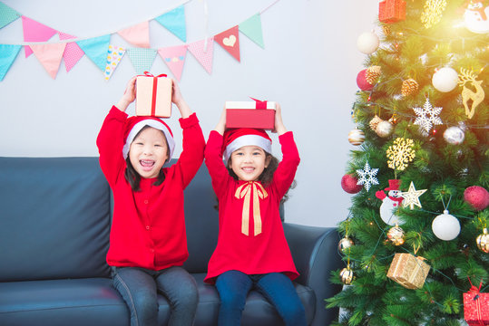 Two little asian girls wearing santa claus hat and holding gift box with smiles beside christmas tree.