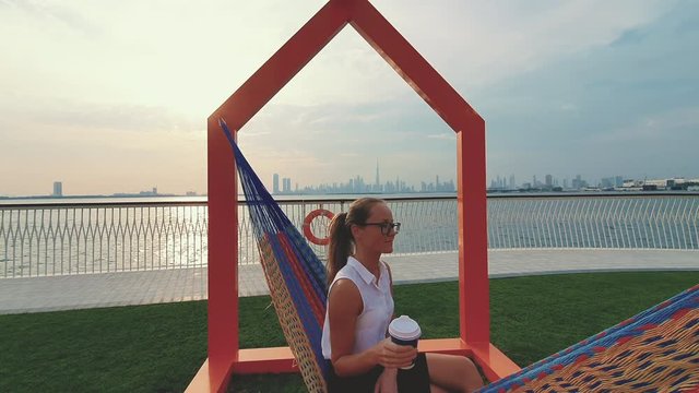 Young woman weared in office sute having coffee in hammock while chilling , freelance life style conceptual, work anywhere. Modern city on the background.