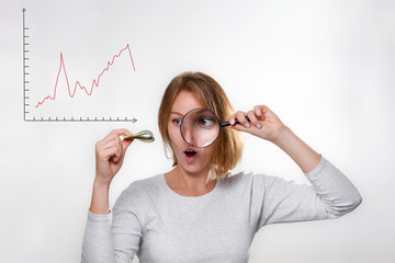 Finance and banking. A young blonde holds a wad of dollars in her hands and looks at them with surprise through a magnifying glass. White background with chart. Copy space