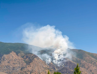 Mountain landscape with smoke from wild forest fire against clear blue sky