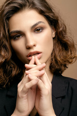 closeup portrait of beauty fashion model with clean skin and curly hair in black jacket on biege background, hands near lips