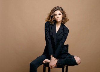 beauty fashion model with clean skin and curly hair in black jacket on biege background on the chair, serious business woman