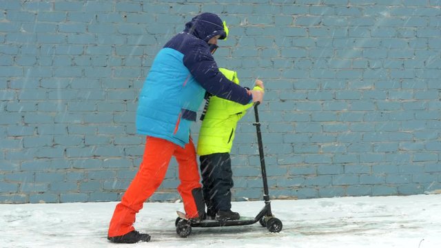 Dad and child in colorful winter clothes and Superman mask riding scooter along gray brick wall in snow. Man and boy waving hands depicting flight