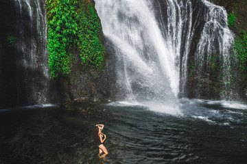 Young slim brunette woman with curly hair enjoying in lagoon of huge tropical waterfall Banyumala...