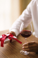 Man's hands with a glass of wine and a gift. Present with a red ribbon.  male hands hold  surprise gift box. Lovers give each other gifts. Valentine's day concept. Romantic day. Birthday anniversary.