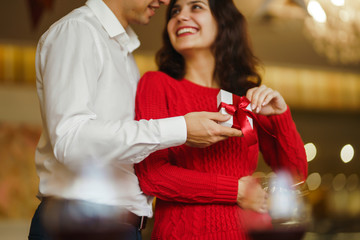 Valentine's Day, holiday and surprise concept. Man gives to his woman a gift box with red ribbon. A loving couple cuddles and celebrating Valentine's Day in the restaurant. Lovers give each other gift