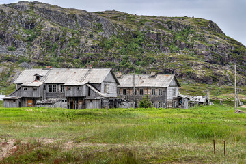 Fototapeta na wymiar Russia, Arctic, Kola Peninsula, Barents Sea, Teriberka: Run down abandoned wooden houses in the city center of the old Russian settlement small fishing village with green grass and grey cloudy sky.