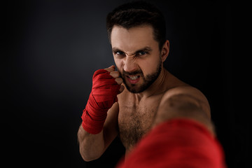 Sportsman boxer throwing a fierce and powerful punch forward in the camera. muscular man with red bandage on hands on black background.