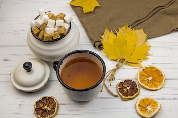 tea in an aluminum cup and sugar in cubes in an aluminum sugar bowl, brown scarf, maple leaves, dried oranges and dried lemon on a light wooden background