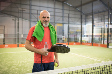 Mature man in paddle tennis court with towel and bottle of water.