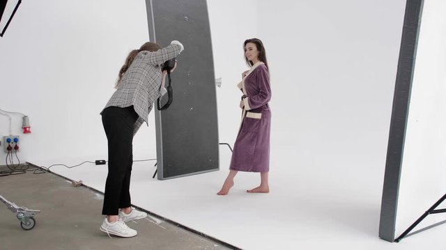 Side view of woman with photo camera shooting barefoot lady in warm bathrobe during photoshoot in professional studio