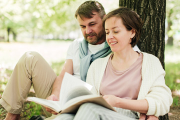 Middle-aged couple sit under a tree reading a book.