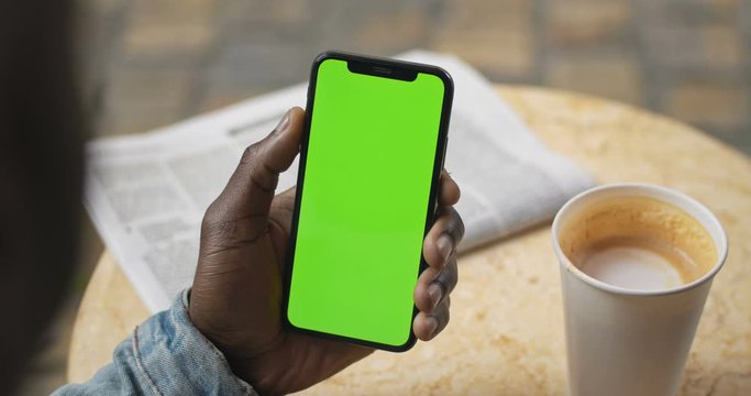 Lviv, Ukraine - August 09, 2019: Hands of Afro American Man Sitting at Table with News Paper and Coffee Cup on it, Holding Smartphone with Green Screen and Touching Screen. Backside View. Close Up.