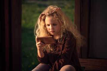 girl child curly-haired blonde sitting on the porch of the house with a mobile phone in her hands, playing games on her mobile