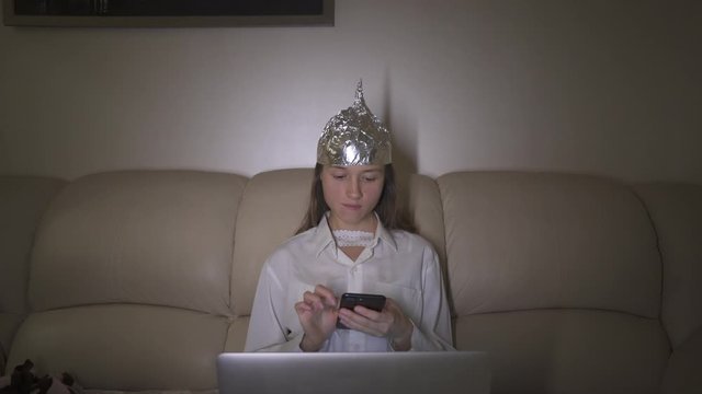 Tin Foil Hat protects woman`s brain from 5G waves, electromagnetic fields, mind control. Girl typing message in mobile phone.