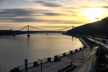 Fototapeta na wymiar Sunrise over Dnipro Dnieper River embankment in Kyiv. Pedestrian bridge on the background. Kyiv, Ukraine. December 2019