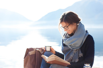 Young woman reading book at sea beach. Cozy winter picnic by morning mountains. Happy student in...