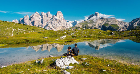 Fototapeta na wymiar Awesome nature Landscape. Unknown couple sitting on green grass over amazing lake with turquoise water of in Tre Cime di Lavaredo national park. Wonderful Dolomites alps scenery.
