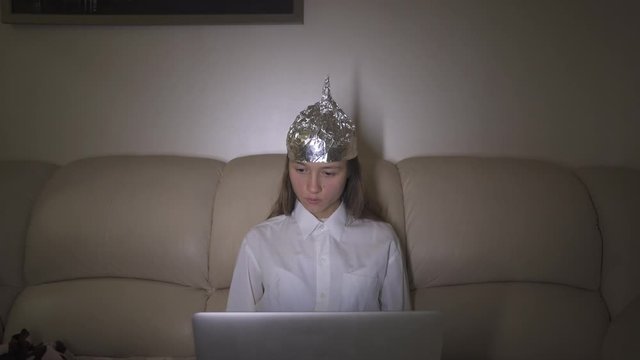 Woman in tin foil hat puts down phone and works at computer, typing on keyboard. Foil hat shields her from 5G waves, internet, electromagnetic fields, mind control and mind reading.